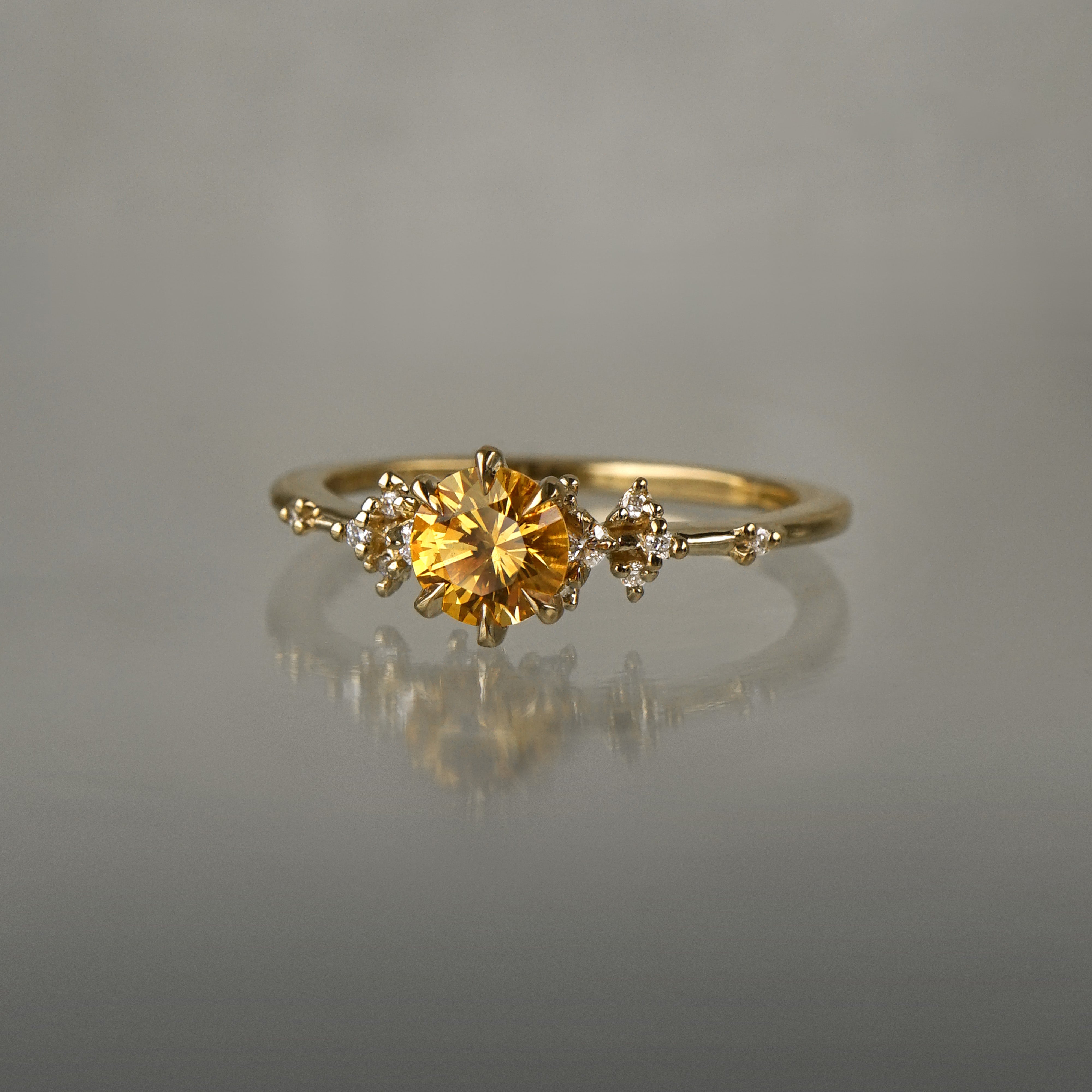 A one of a kind 18k yellow gold Water Lily engagement ring by Laurie Fleming Jewellery with a bright golden yellow round Montana sapphire centre with clusters of diamonds on the band. The ring is situated on a medium grey background.