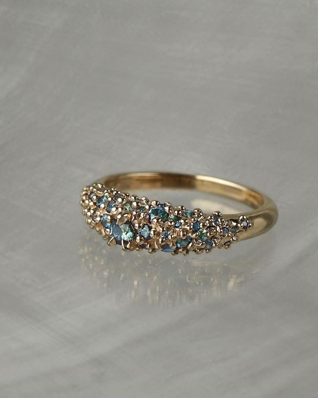 Laurie Fleming Jewellery's "Moss Signet Ring," a bold solid gold signet-style ring encrusted with varying shades and sizes of blue-green teal sapphires on the band. The ring is resting on a grey reflective surface.