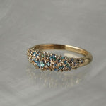 Laurie Fleming Jewellery's "Moss Signet Ring," a bold solid gold signet-style ring encrusted with varying shades and sizes of blue-green teal sapphires on the band. The ring is resting on a grey reflective surface.