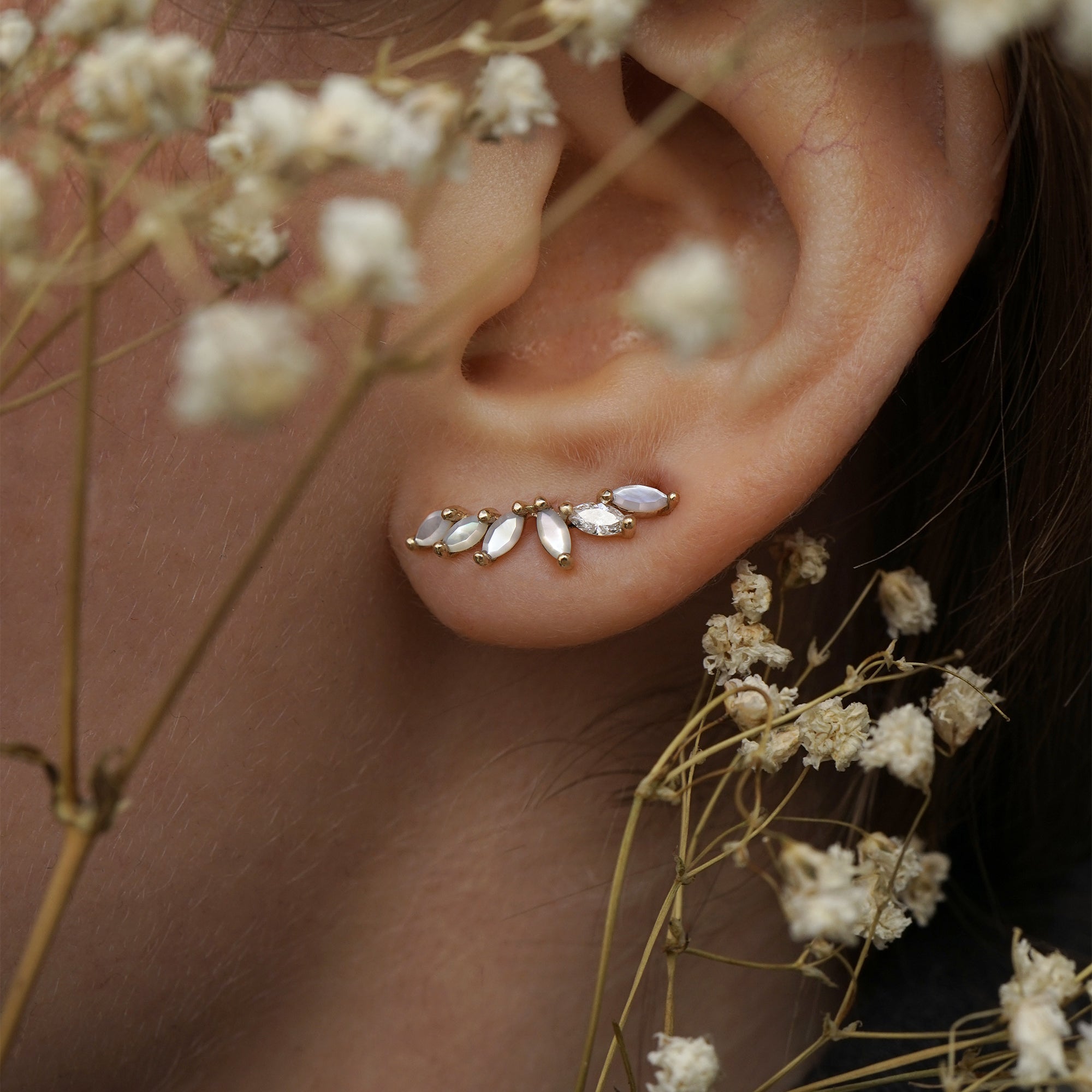 A close up of an ear wearing a Lyrie stud earring with marquise cut mother of pearl and a single marquise cut diamond. There are dried flower stems in the foreground around the ear.