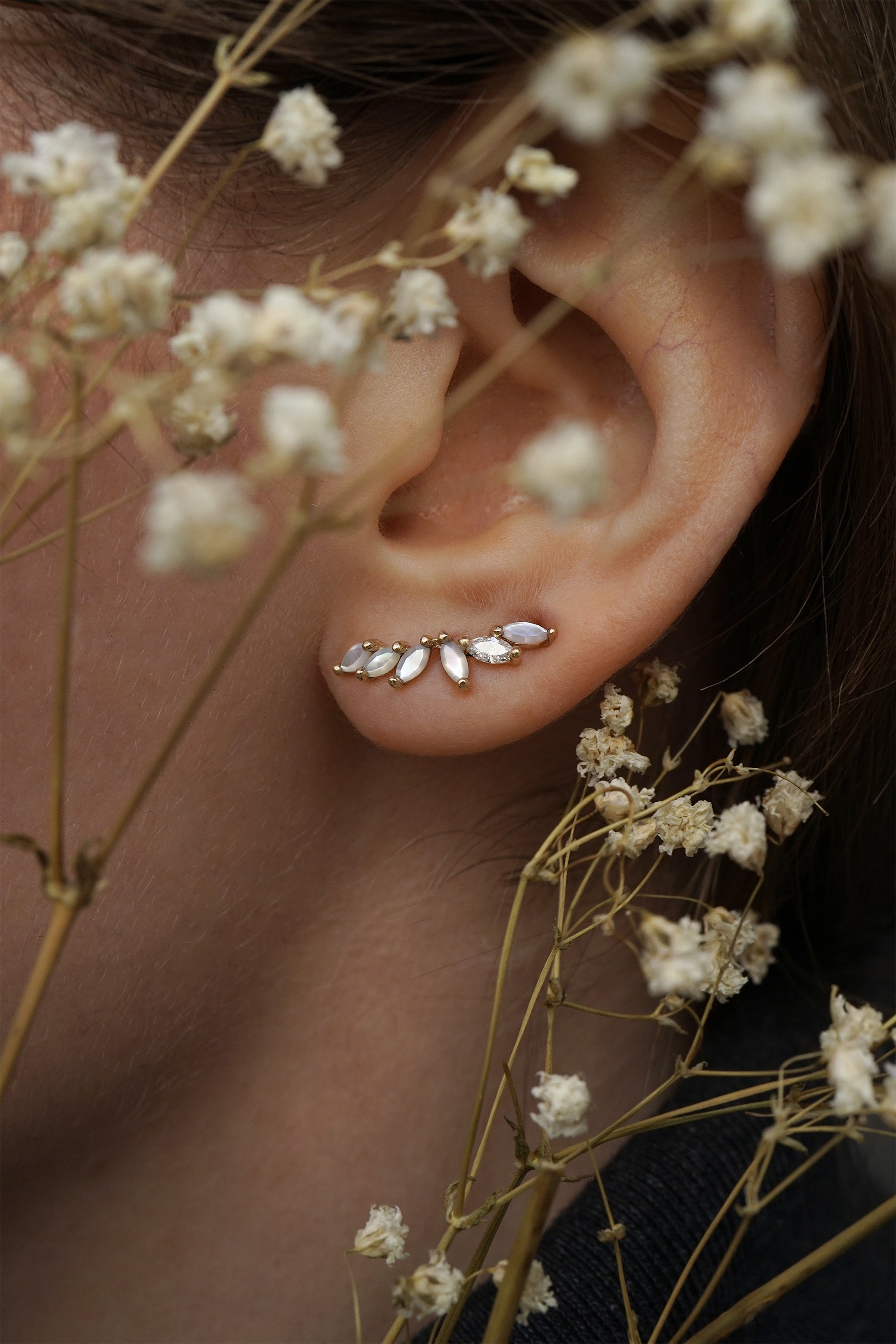 A close up of an ear wearing a Lyrie stud earring with marquise cut mother of pearl and a single marquise cut diamond. There are dried flower stems in the foreground around the ear.