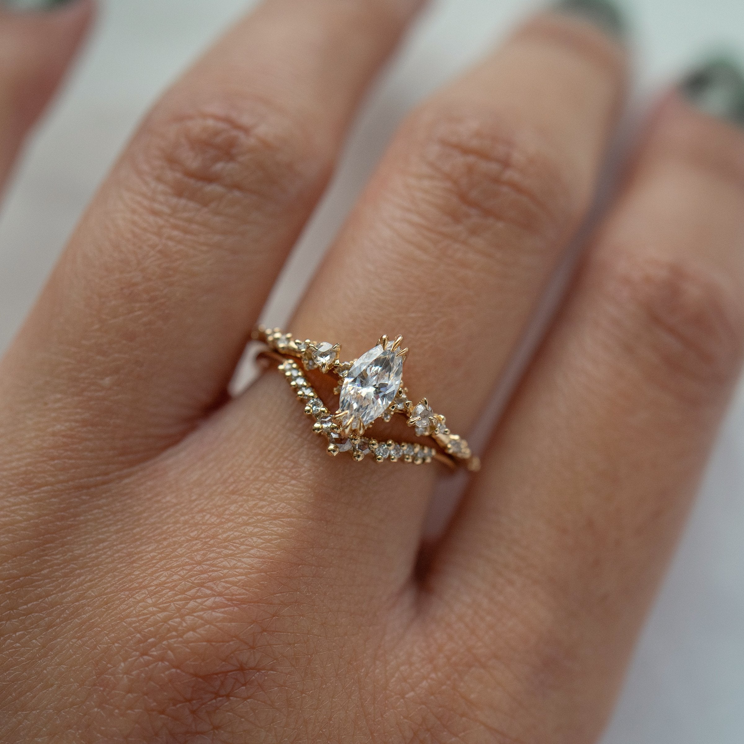 A close up of a hand wearing two solid gold rings by Laurie Fleming Jewellery: on the top, the Nereid marquise cut diamond engagement ring, and on the bottom, the Selene contour band ring, which features rose cut and brilliant cut diamonds along a curve to stack with other rings (such as with an engagement ring).