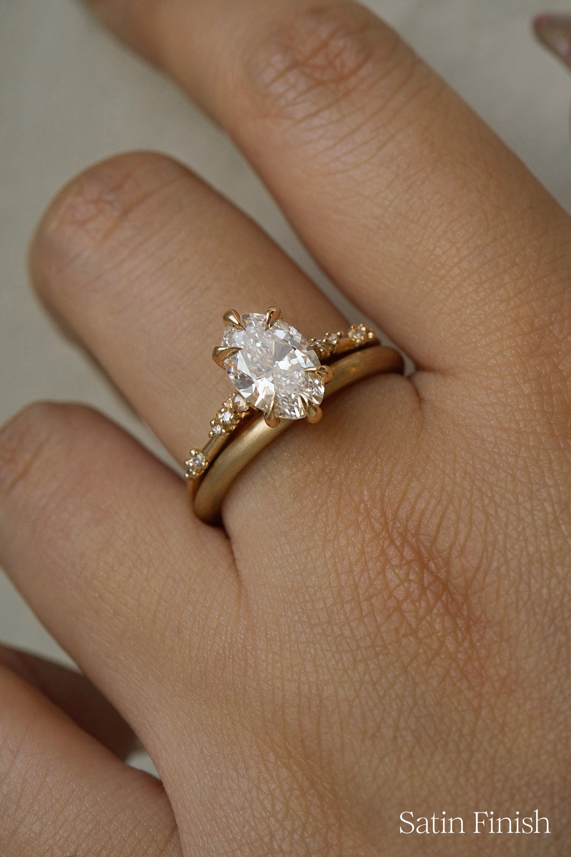 A photo of a hand wearing two gold rings by Laurie Fleming Jewellery in front of a light grey background. On top is a one of a kind Daphne ring with an oval cut diamond centre and diamonds on the band. On the bottom, a plain round "Peach" wedding band stacking ring, with a soft satin brushed finish.