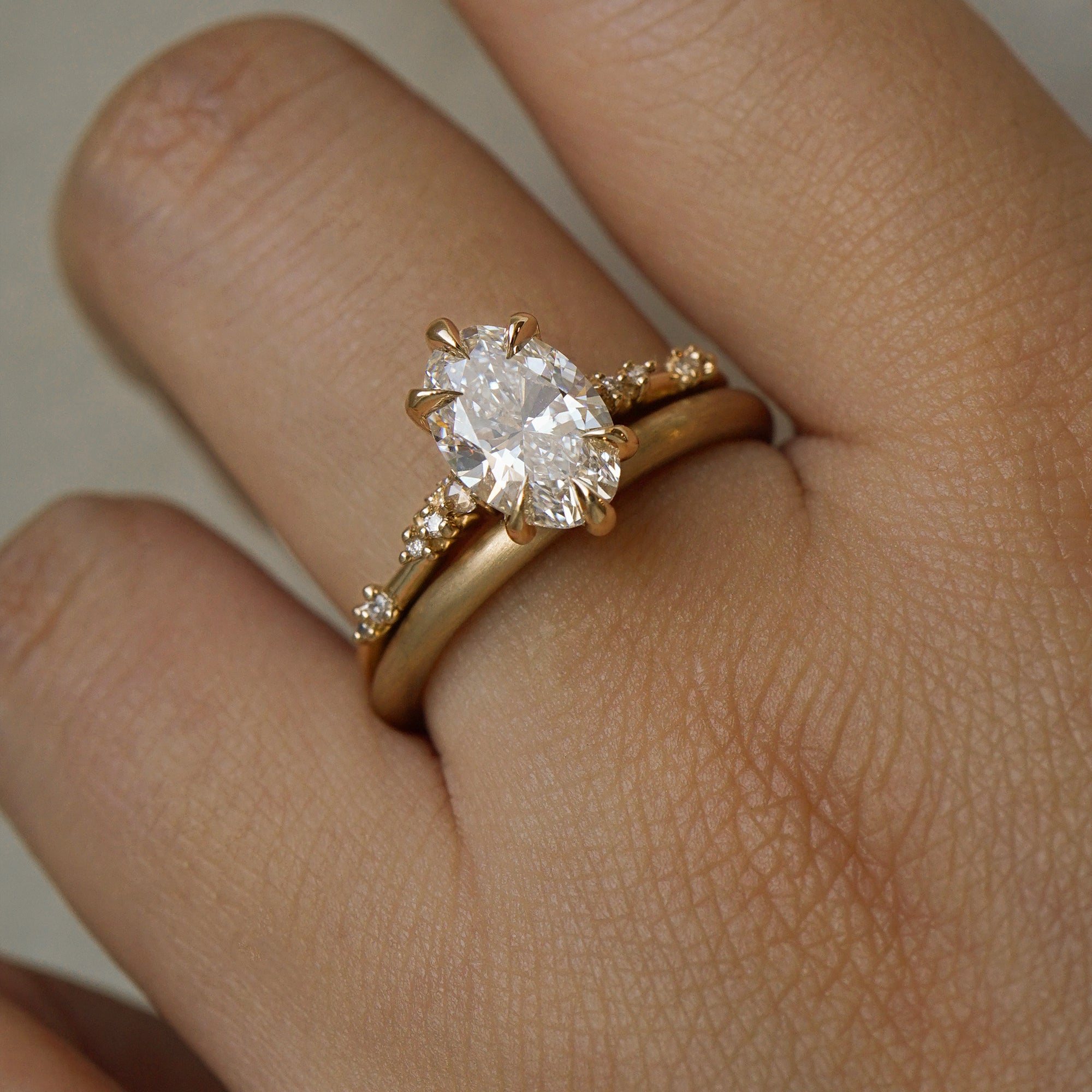A photo of a hand wearing two gold rings by Laurie Fleming Jewellery in front of a light grey background. On top is a one of a kind Daphne ring with an oval cut diamond centre and diamonds on the band. On the bottom, a plain round "Peach" wedding band stacking ring, with a soft satin brushed finish.