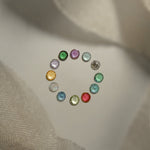 A photo of the different birthstone gems that are available in the Laurie Fleming Jewellery "Pansy" collection, arranged in a ring on a a light grey background with bits of silk at the top and bottom of the frame. From the top of the circle, clockwise:  Amethyst, Aquamarine, Diamond, Emerald, Alexandrite, Ruby, Peridot, Blue Sapphire, Australian Opal, Citrine, Tanzanite, Tsavorite Garnet.