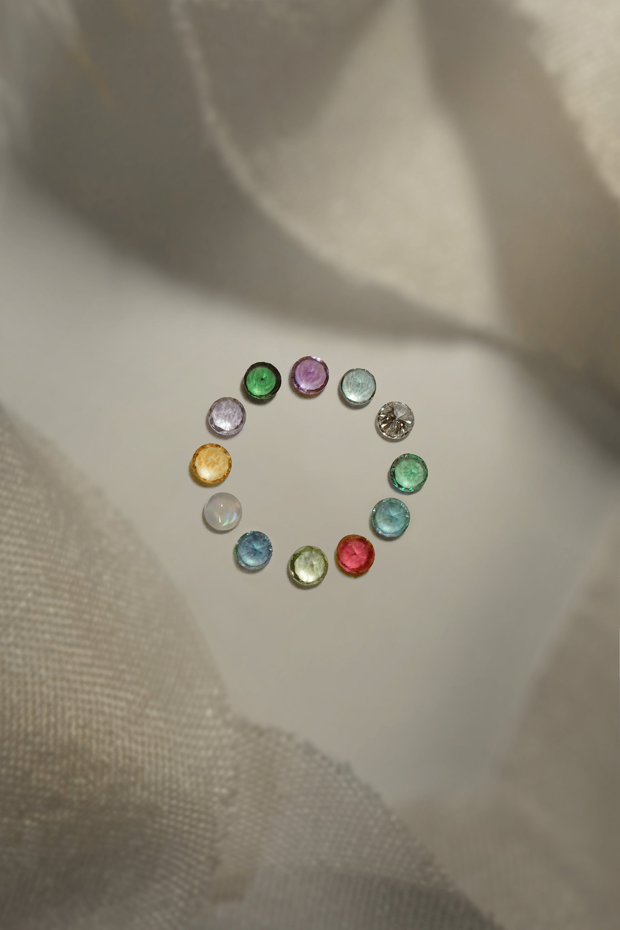 A photo of the different birthstone gems that are available in the Laurie Fleming Jewellery "Pansy" collection, arranged in a ring on a a light grey background with bits of silk at the top and bottom of the frame. From the top of the circle, clockwise:  Amethyst, Aquamarine, Diamond, Emerald, Alexandrite, Ruby, Peridot, Blue Sapphire, Australian Opal, Citrine, Tanzanite, Tsavorite Garnet.