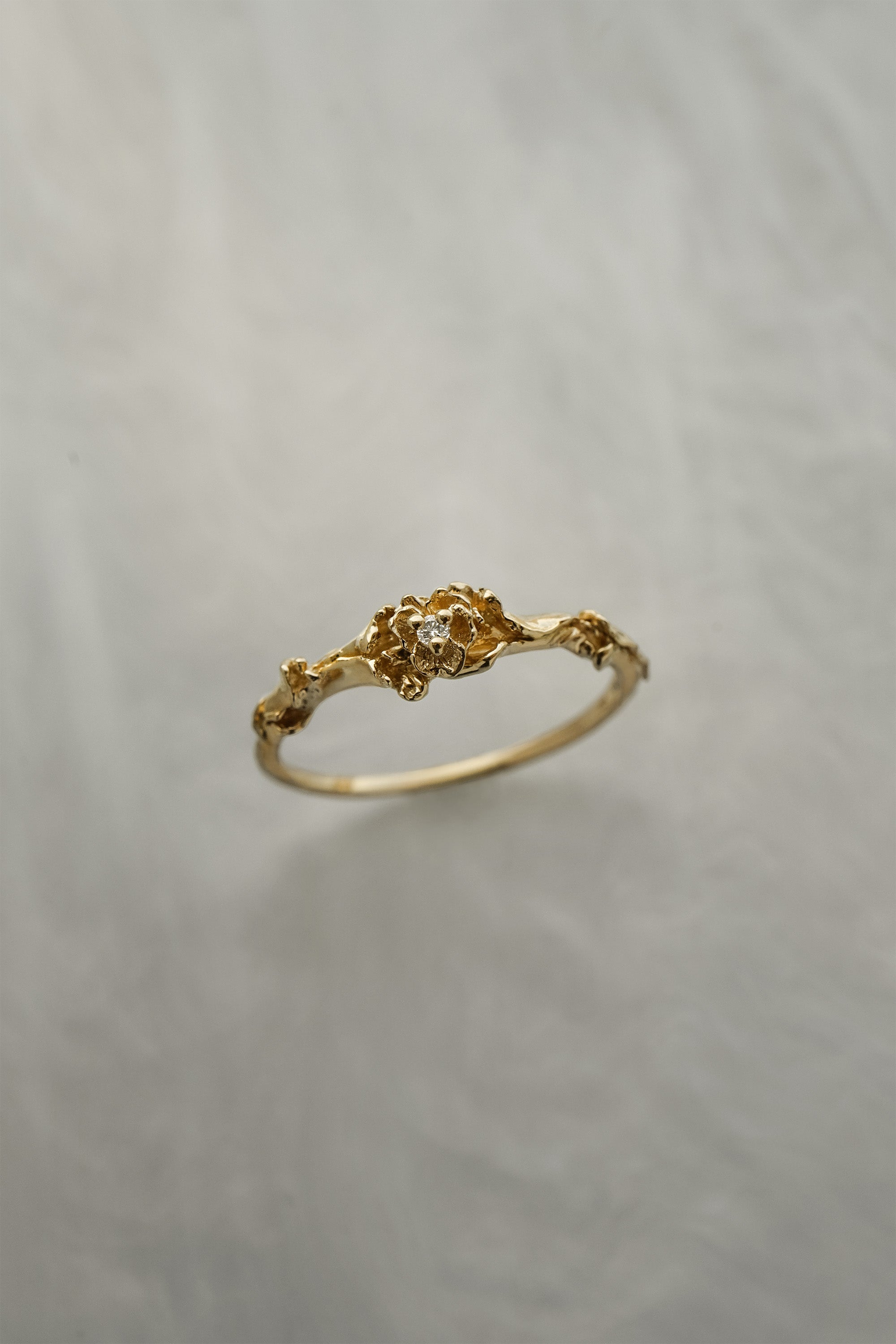 A stock photo of the solid gold "Pansy Signet" ring by Laurie Fleming Jewellery. The ring features hand-carved delicate petals encircling the band, with a tiny pansy flower at the centre, which has a customizable centre stone (pictured with a diamond, available in all birthstones). The ring is against a medium/light grey background.