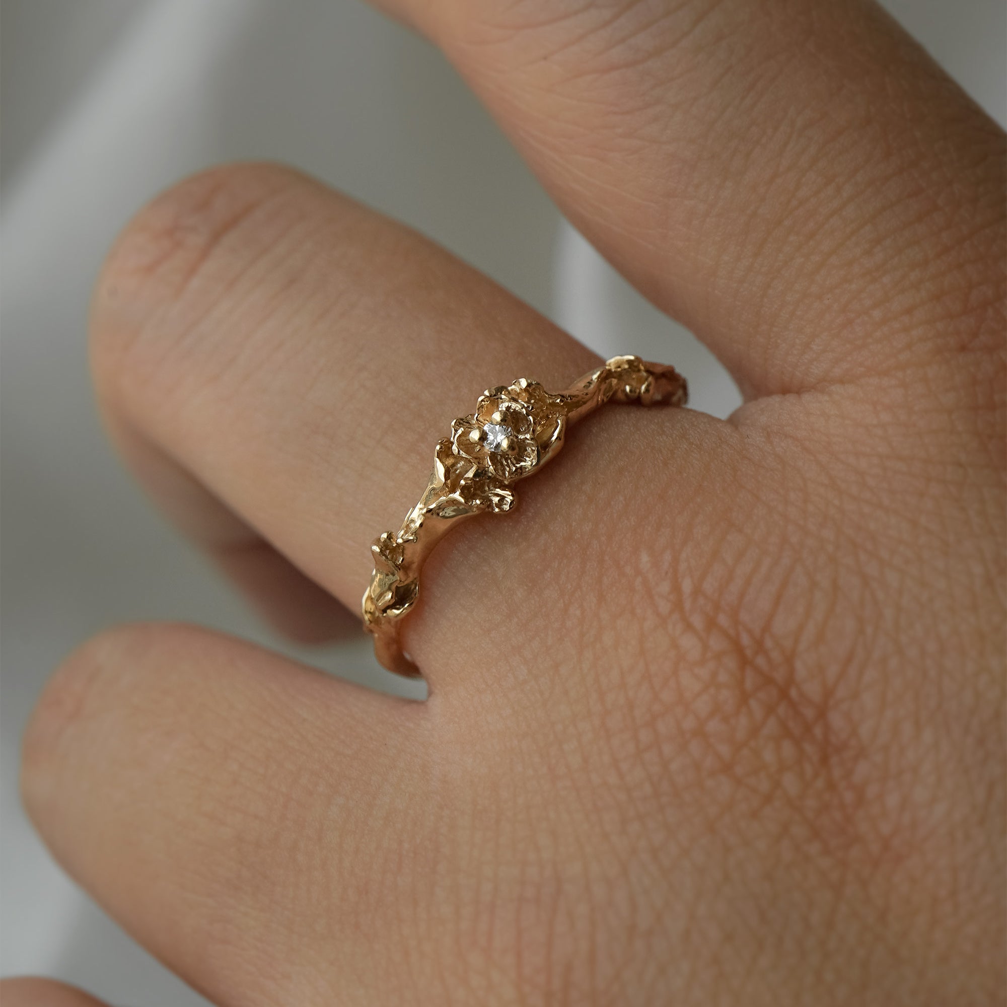 A stock photo of the solid gold "Pansy Signet" ring by Laurie Fleming Jewellery. The ring features hand-carved delicate petals encircling the band, with a tiny pansy flower at the centre, which has a customizable centre stone (pictured with a diamond, available in all birthstones). The ring is worn on a hand against a light grey background.