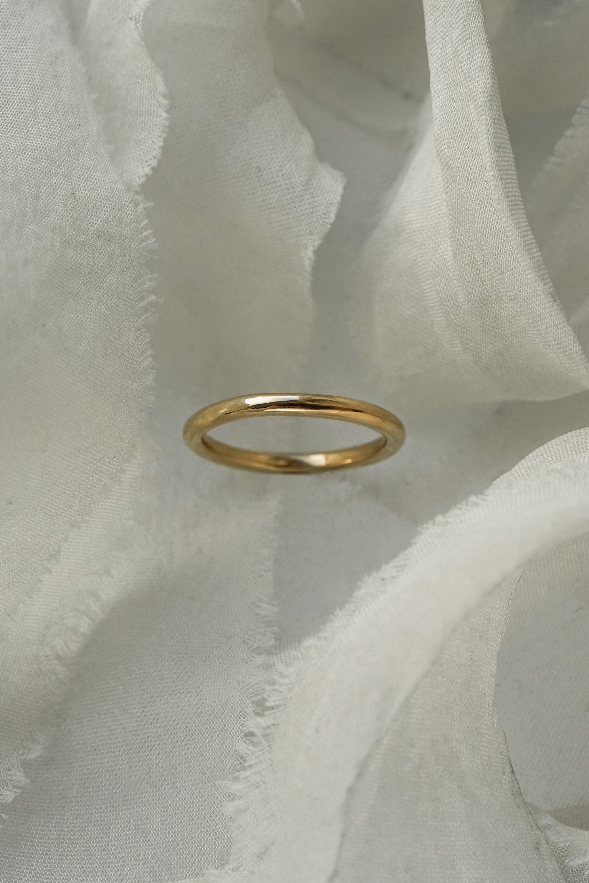 A stock photo of Laurie Fleming Jewellery's plain solid gold "Peach" band, a 2.1mm wide round wire stacking ring/wedding band, handmade in Toronto. The ring is standing on its own surrounded by pieces of raw white silk.