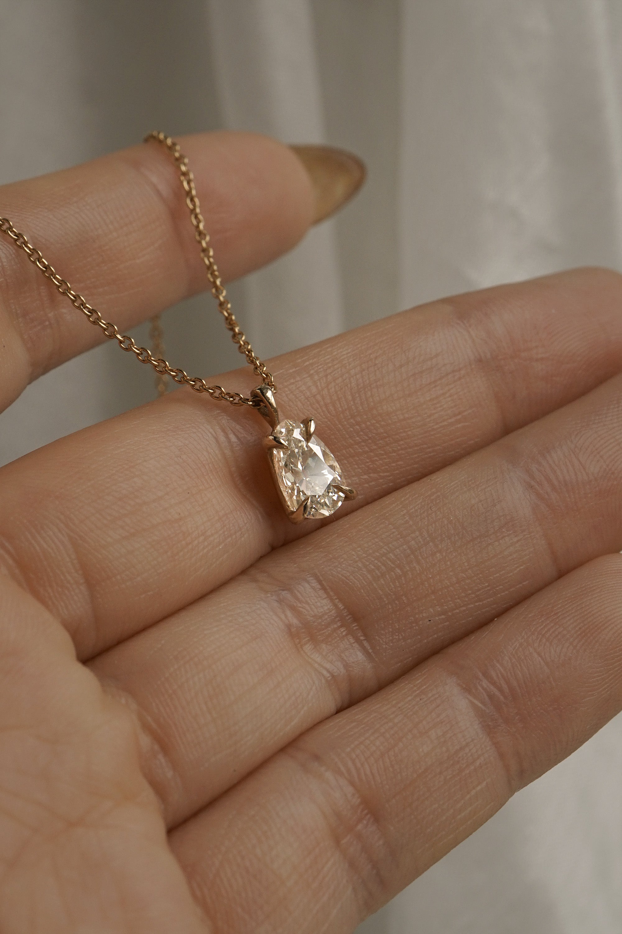 A hand holding a one of a kind, handcarved solid 14k yellow gold necklace featuring a 1.07ct antique pear cut champagne diamond. The diamond is held in four delicate claw-shaped prongs. The pendant hangs from an adjustable 16-18" chain, and is draped across the hand's fingers. In the background, some white gossamer silk fabric hangs.