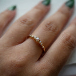 A hand in front of a light grey background wearing a Laurie Fleming Jewellery "Pearl Water Lily Ring," a delicate solid gold stacking ring with freshwater seed pearls scattered along the band. 