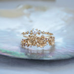 A stack of dainty Laurie Fleming Jewellery rings. The top ring is a "Pearl Water Lily Ring," a delicate solid gold stacking ring with freshwater seed pearls scattered along the band. The bottom ring is an "Asrai Garden" style ring, featuring three handcarved miniature orchid flowers with freshwater seed pearls, and lilac with pink and purple sapphires. Rings are stacked on a pearlescent oyster dish.