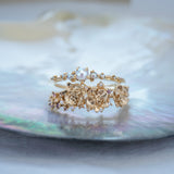 A stack of dainty Laurie Fleming Jewellery rings. The top ring is a "Pearl Water Lily Ring," a delicate solid gold stacking ring with freshwater seed pearls scattered along the band. The bottom ring is an "Asrai Garden" style ring, featuring three handcarved miniature orchid flowers with freshwater seed pearls, and lilac with pink and purple sapphires. Rings are stacked on a pearlescent oyster dish.