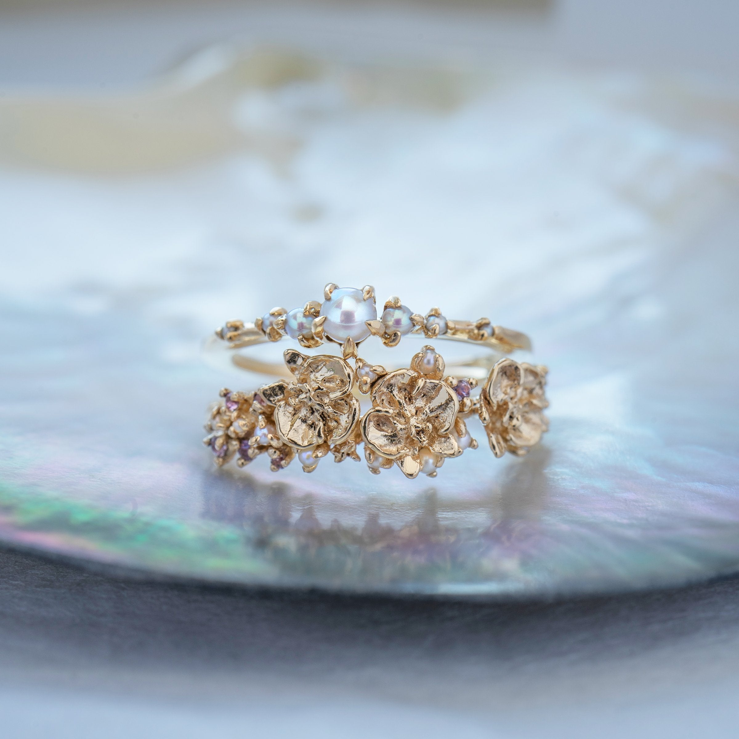 A stack of dainty Laurie Fleming Jewellery rings. The top ring is a "Pearl Water Lily Ring," a delicate solid gold stacking ring with freshwater seed pearls scattered along the band. The bottom ring is an "Asrai Garden" style ring, featuring three handcarved miniature orchid flowers with freshwater seed pearls, and lilac with pink and purple sapphires. Rings are stacked on a pearlescent oyster dish.