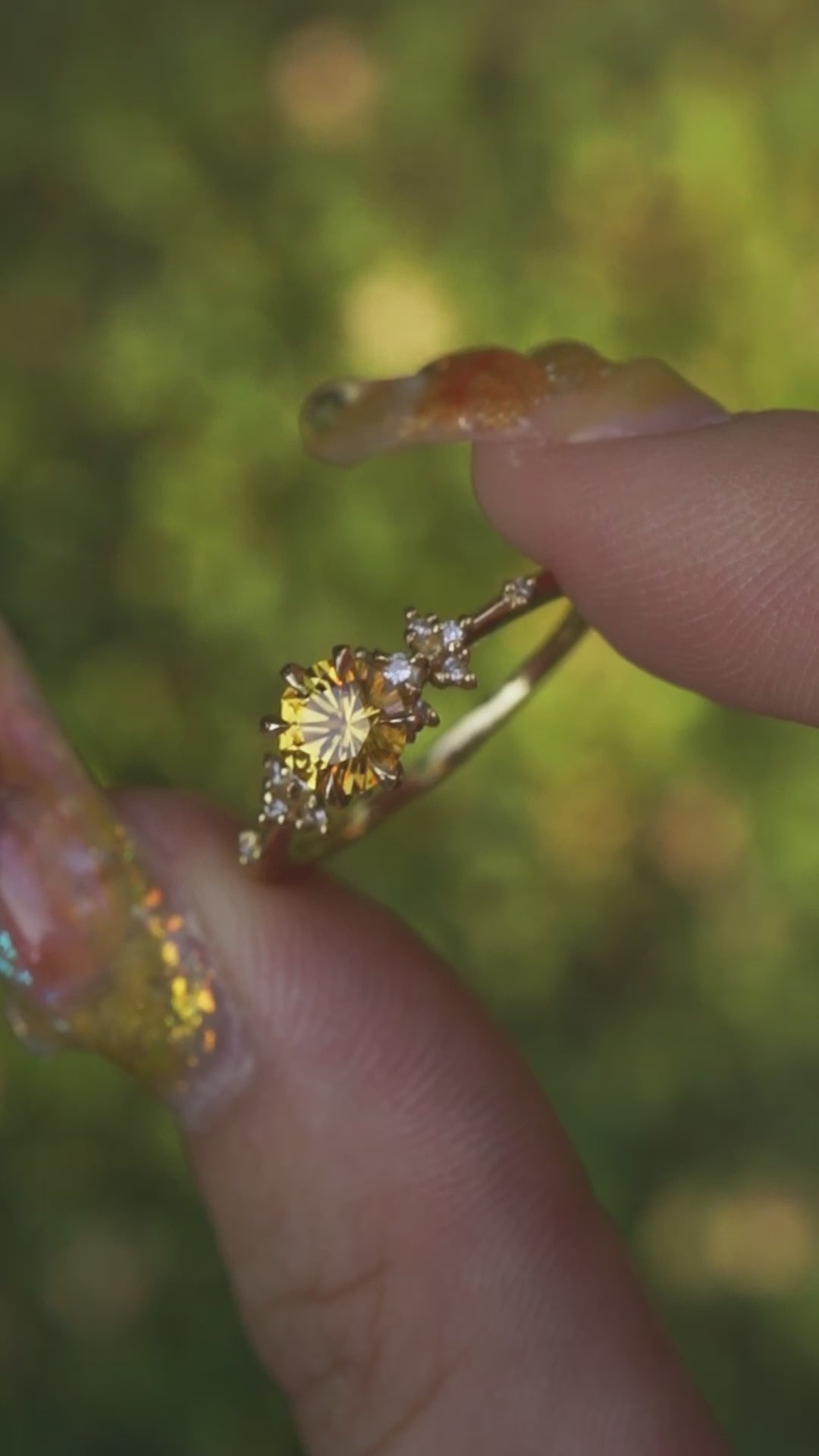 A one of a kind 18k yellow gold Water Lily engagement ring by Laurie Fleming Jewellery with a bright yellow round Montana sapphire centre with clusters of diamonds on the band. The ring is held between index finger and thumb and gently rotated before a background of unfocused plants in shades of soft green.