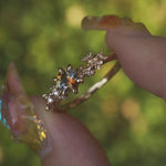 A one of a kind "Asai Garden" style engagement ring by Laurie Fleming Jewellery with a round orange-blue "Kaleidoscope" Montana sapphire centre. The band features hand carved cherry blossom flowers with brilliant diamonds. The ring is held between an index finger and thumb and rotated gently in front of a background of soft unfocused greenery.