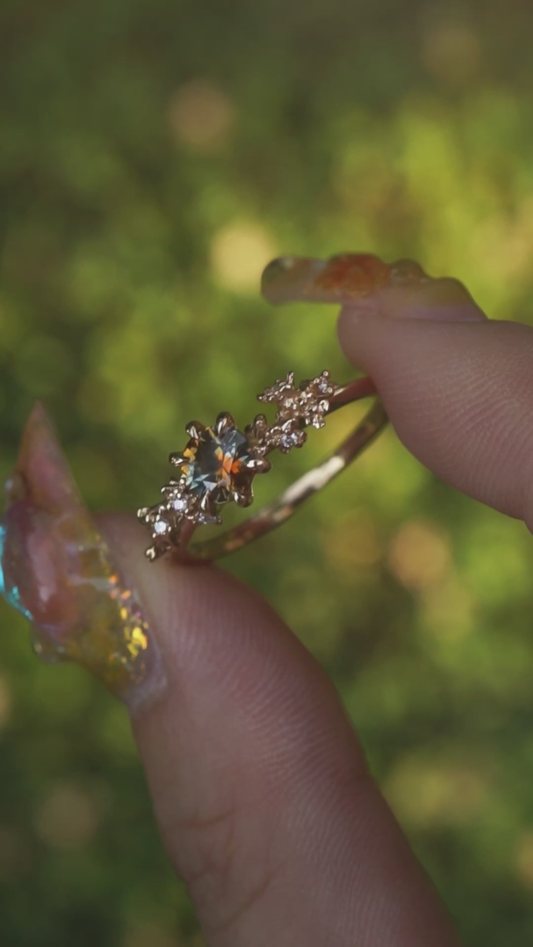 A one of a kind "Asai Garden" style engagement ring by Laurie Fleming Jewellery with a round orange-blue "Kaleidoscope" Montana sapphire centre. The band features hand carved cherry blossom flowers with brilliant diamonds. The ring is held between an index finger and thumb and rotated gently in front of a background of soft unfocused greenery.