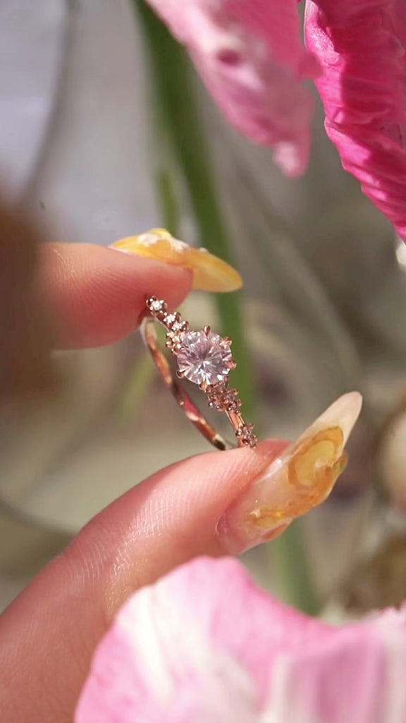 A one of a kind "Daphne" style engagement ring by Laurie Fleming Jewellery,held between index finger and thumb and rotated side to side.  The ring features a pale rosewater pink sapphire centre. The band is encrusted in sparkling diamond clusters. The ring is handcarved and made in solid champagne yellow gold. There are partially visible flowers in the foreground and the background, with the focus on the ring at the centre of the frame.