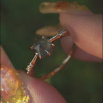 A dainty yellow gold Ilona style engagement ring with a rose cut green sapphire and diamonds on the band, held between index finger and thumb and rotated gently over a background of soft unfocused greenery.