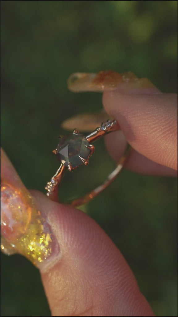 A dainty yellow gold Ilona style engagement ring with a rose cut green sapphire and diamonds on the band, held between index finger and thumb and rotated gently over a background of soft unfocused greenery.