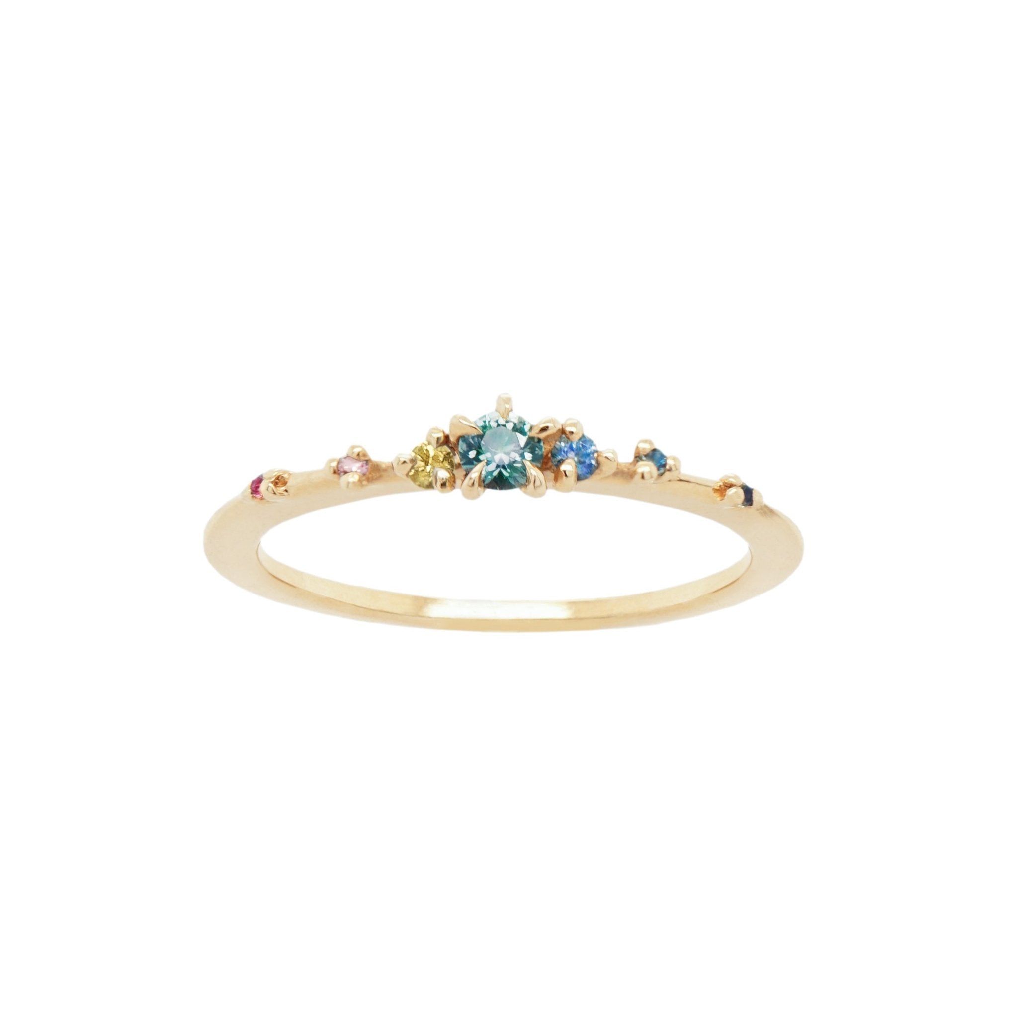 Laurie Fleming Jewellery's "Rainbow Sapphire Water Lily Ring," a delicate solid gold stacking ring with a rainbow gradient of sapphires (deep pink sapphire, light pink sapphire, yellow sapphire, teal sapphire, light blue sapphire, medium blue sapphire, dark blue sapphire) scattered along the band. The ring is on a white background.