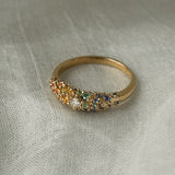 Laurie Fleming Jewellery's "Rainbow Sapphire Moss Signet Ring," a bold solid gold signet-style ring encrusted with sapphires in varying shades and sizes in deep pink, medium pink, light pink, orange, yellow, green, blue-green, medium blue, light blue, and purple, with a single round brilliant cut diamond at the centre on the band. The ring is resting on white gossamer silk.