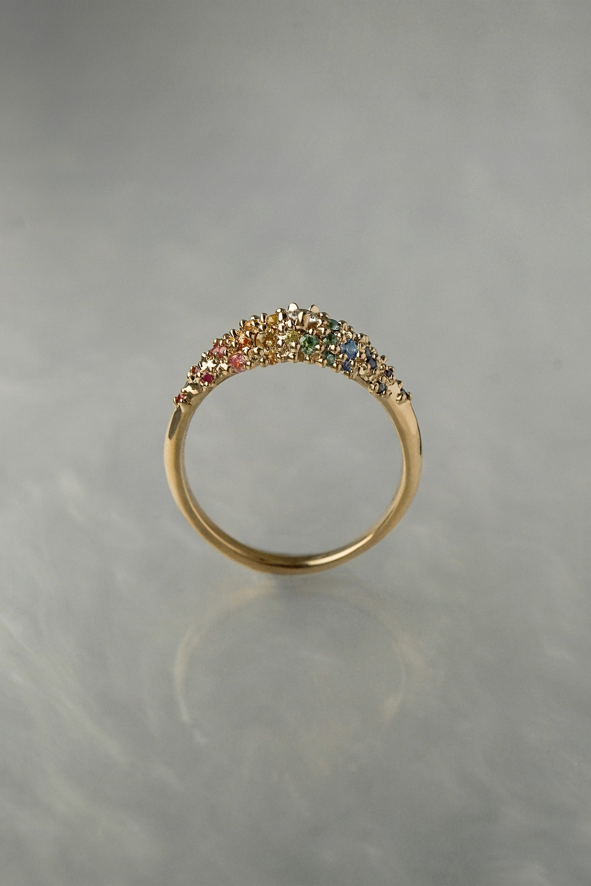 Laurie Fleming Jewellery's "Rainbow Sapphire Moss Signet Ring," a bold solid gold signet-style ring encrusted with sapphires in varying shades and sizes in deep pink, medium pink, light pink, orange, yellow, green, blue-green, medium blue, light blue, and purple, with a single round brilliant cut diamond at the centre on the band. The ring is seen from a side view in front of a medium grey background.