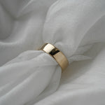 A stock photo of Laurie Fleming Jewellery's plain solid gold "Rowan" band, a 6mm wide flat comfort fit stacking ring/wedding band, handmade in Toronto. The ring is held on a roll of white silk fabric which is nestled against more silk.