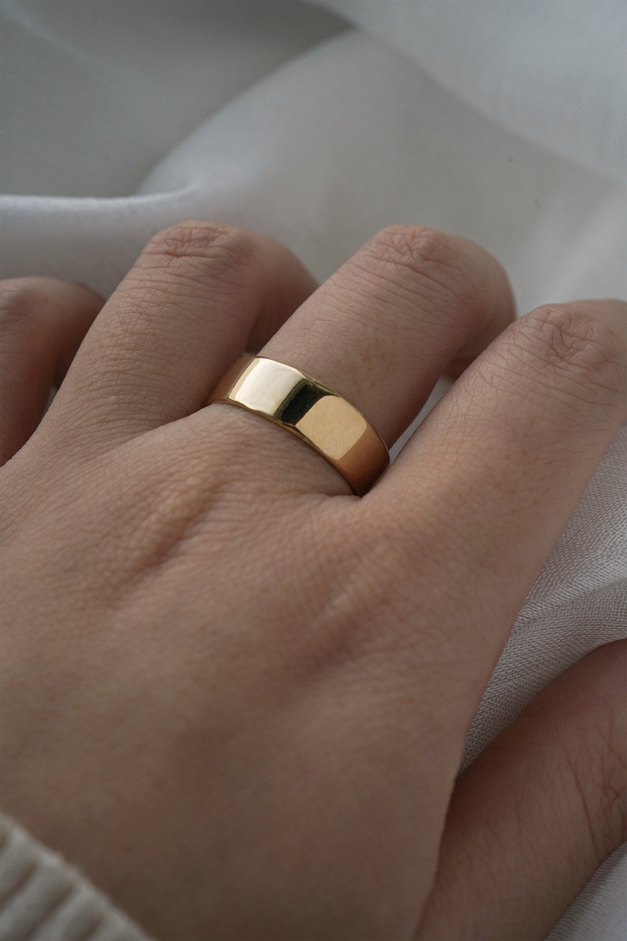 A stock photo of Laurie Fleming Jewellery's plain solid gold "Rowan" band, a 6mm wide flat comfort fit stacking ring/wedding band, handmade in Toronto. The ring is worn on a hand with a light grey background.