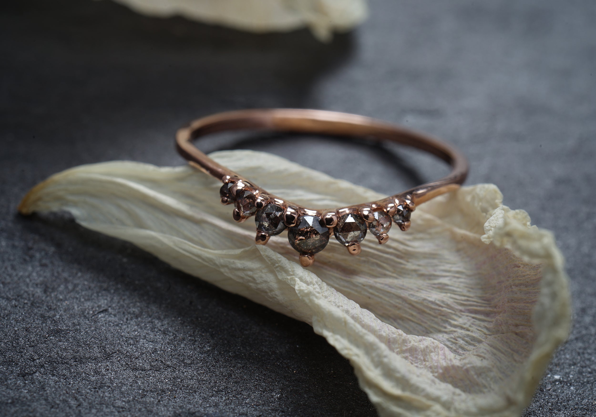 A product photo showing the salt and pepper rose cut diamond Beacen Ring by Laurie Fleming Jewellery. The ring features a gentle tapered curve that stacks well with other rings or contours against engagements rings. It is resting against a dried white flower petal on a grey background.