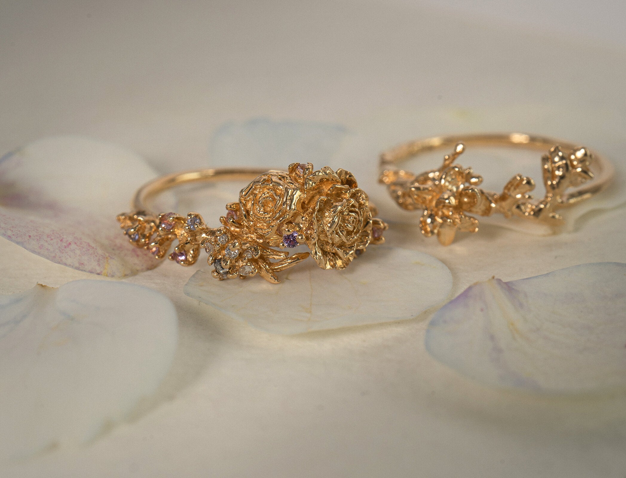 Two one of a kind Asrai Garden Rings. On the right, the ring features Sleeping Petal Band, Anemone, Lilacs, and Ranunculus. On the left, slightly out of focus, the ring features Sleeping Petal Band, Magnolia Branch. - Laurie Fleming Jewellery