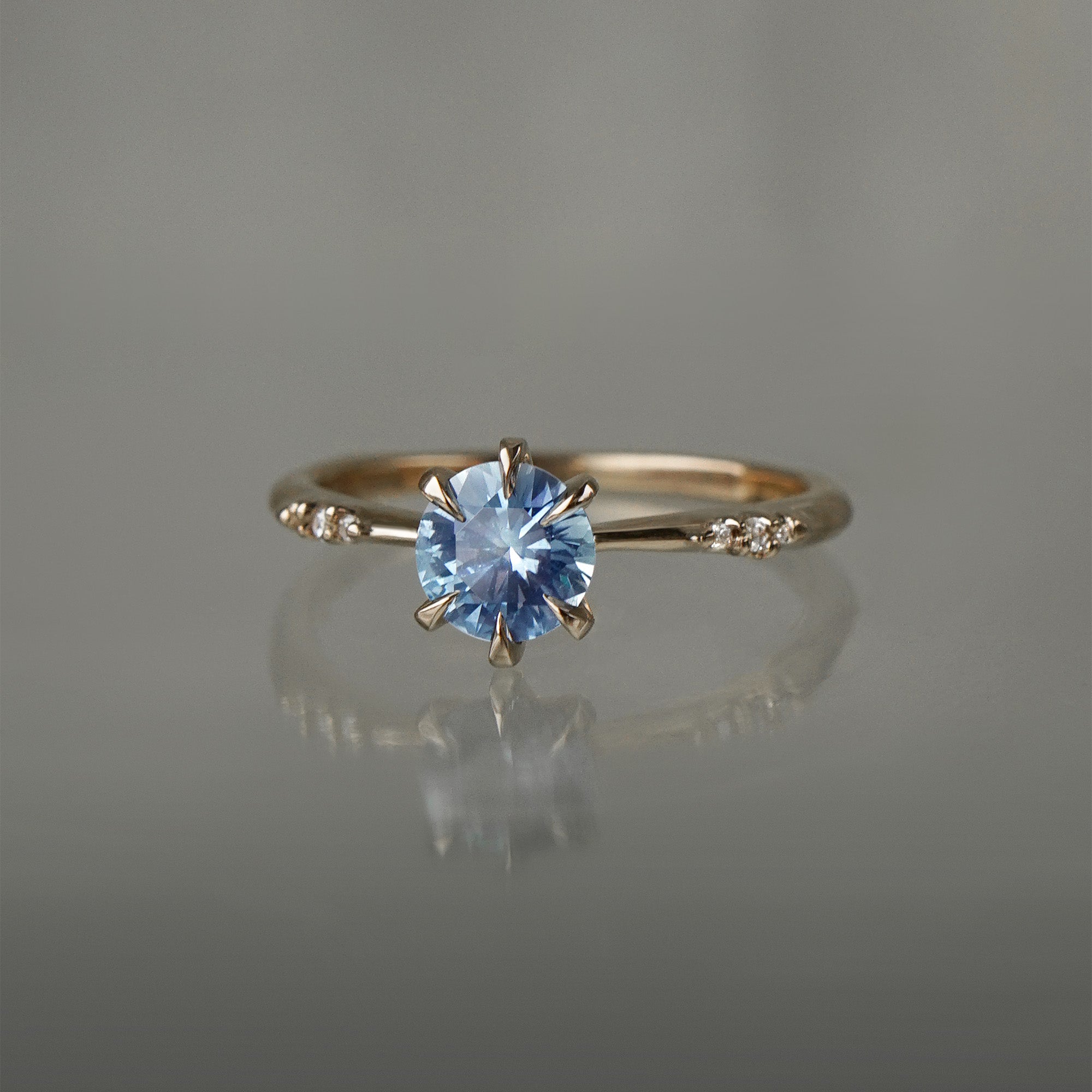 A delicate "Ilona" style one of a kind engagement ring by Laurie Fleming Jewellery in solid 14k champagne gold. It features a stunning sky blue colored round sapphire, held by six hand-shaped claw prongs. The band features small diamonds on the shoulders. The ring is situated on a grey background.