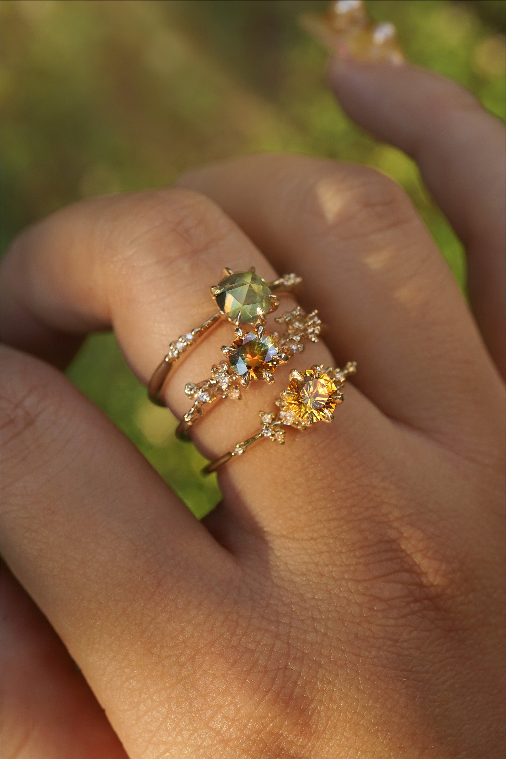 A hand wearing three yellow gold rings by Laurie Fleming Jewellery. The top ring is an Ilona style and features a round rose cut green sapphire on a delicate band with diamonds on the shoulders. In the middle, an Asrai Garden ring with a round orange-blue sapphire centre and carved cherry blossoms on the band featuring diamonds. At the base of the finger, a one of a kind Water Lily ring with a round bright yellow sapphire with clusters of diamonds on the band. The background looks like out of focus plants.