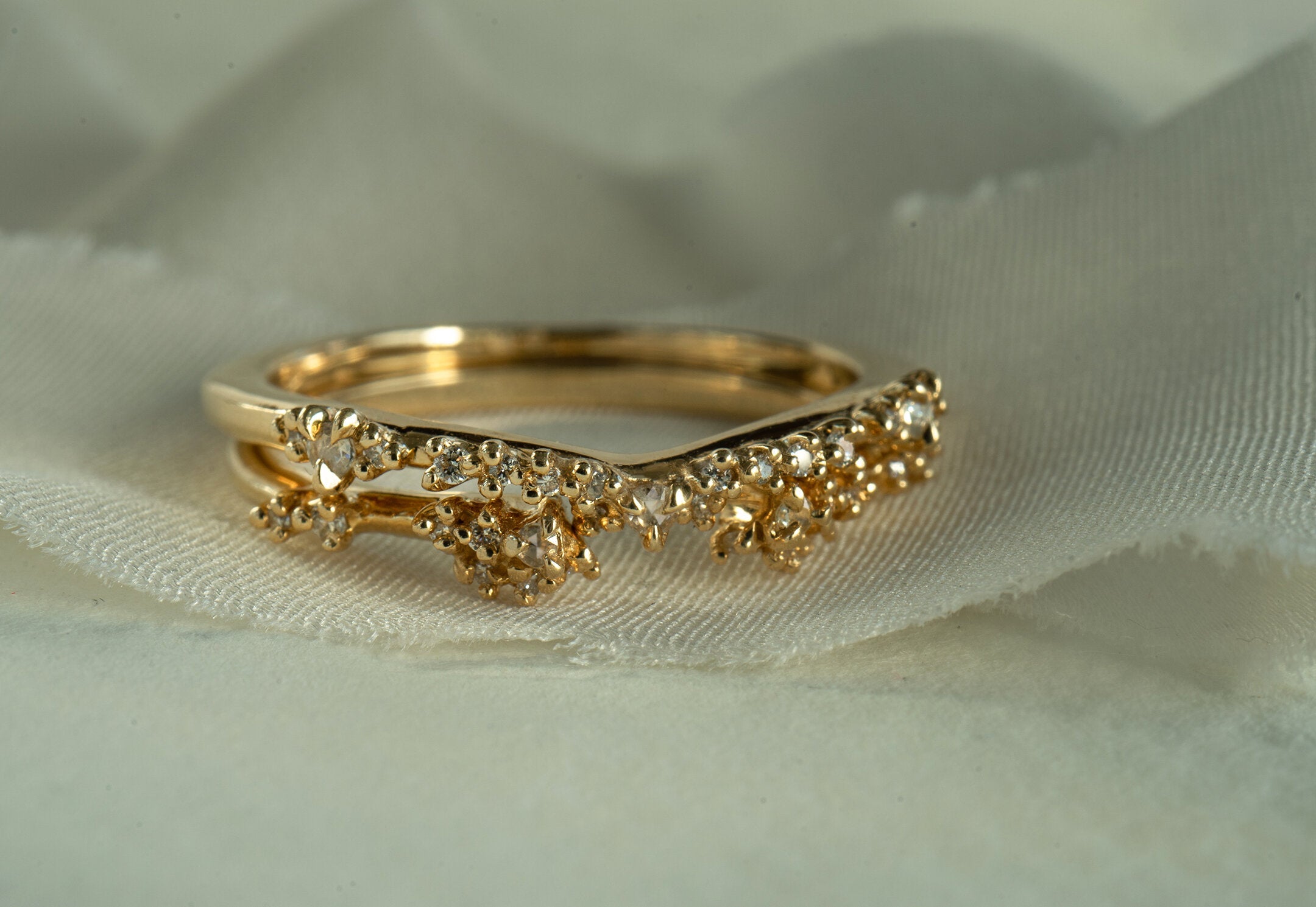 A photo of two Laurie Fleming Jewellery rings stacked together against a background of white fabric. On top, the Sylph band. Below, the Daphne Band Cuff ring. Both rings feature rose cut and brilliant cut diamond accents on the bands.