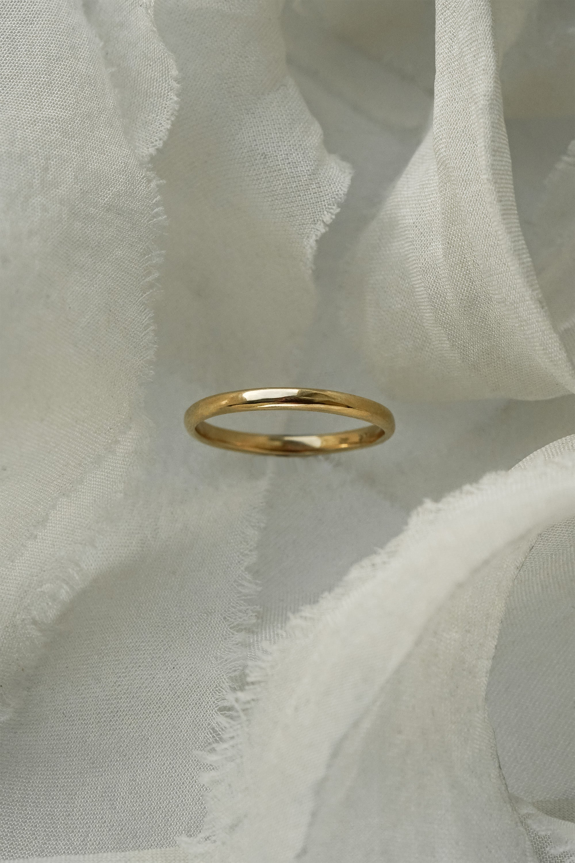 A stock photo of Laurie Fleming Jewellery's plain solid gold "Wisteria" band, a 2mm wide stacking ring/wedding band, handmade in Toronto. The ring is standing on its own surrounded by pieces of raw white silk.