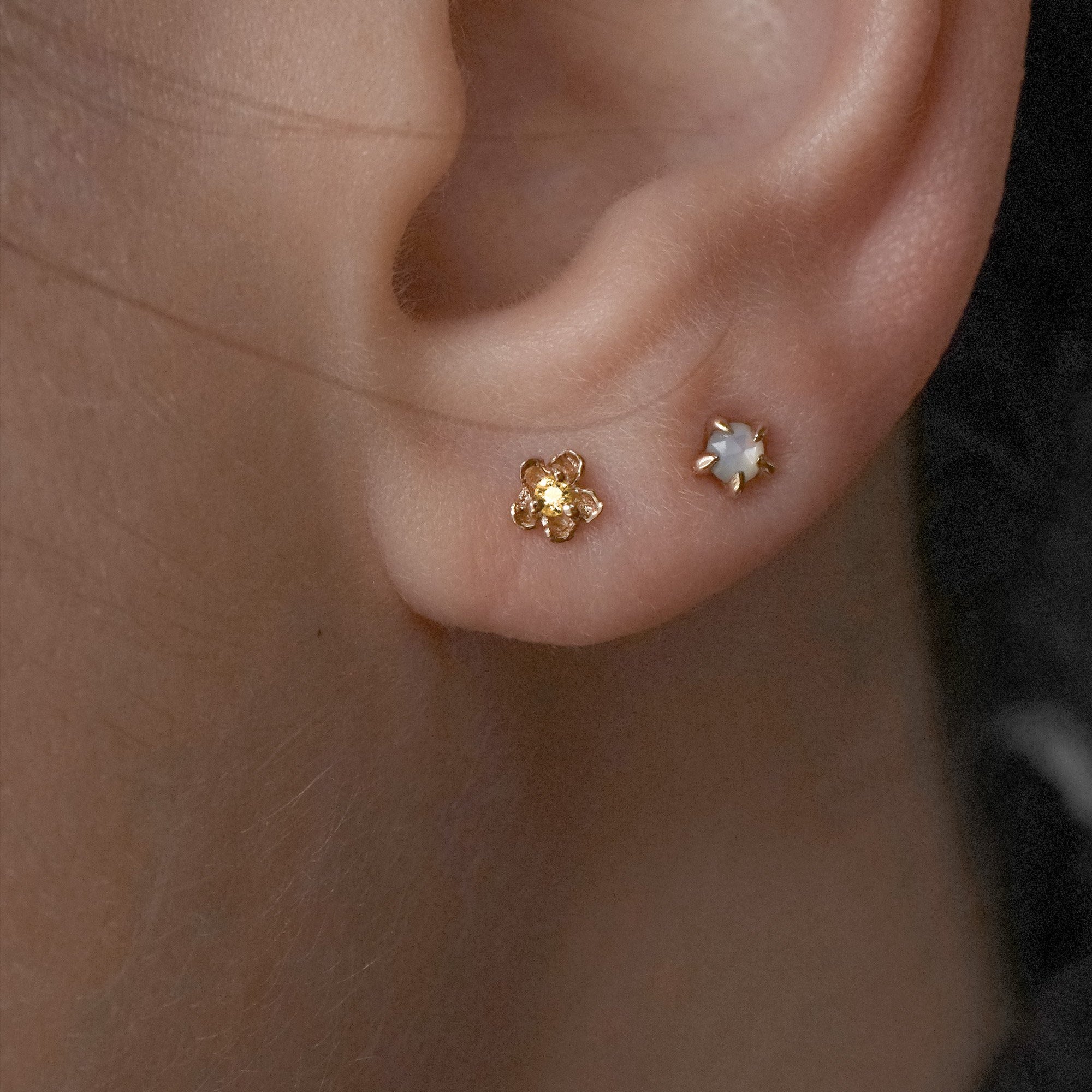 A close-up shot of an ear with two piercings wearing two petite studs by Laurie Fleming Jewellery. In the first hole, a "Buttercup Stud" with a hand-carved buttercup flower with a yellow sapphire, and in the second hole, a "Mother of Pearl Moondew Stud", with a rose cut mother of pearl stone held by five delicate claw-shaped prongs.