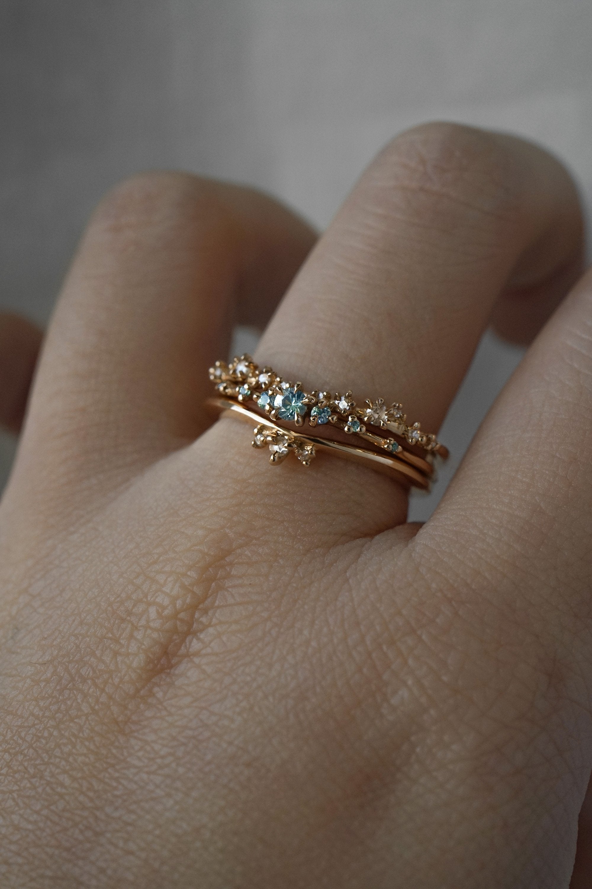 A stack of delicate Laurie Fleming Jewellery gold rings. At the base of the finger, the "Dewdrop Band", a gently curved and tapered band with three rose cut diamonds at the centre on one side. In the middle, a "Sapphire Water Lily Ring," a delicate solid gold stacking ring with blue-green teal sapphires scattered along the band. On top, the "White Wisp Band," a wedding band with scattered rose and brilliant cut diamonds on the band.