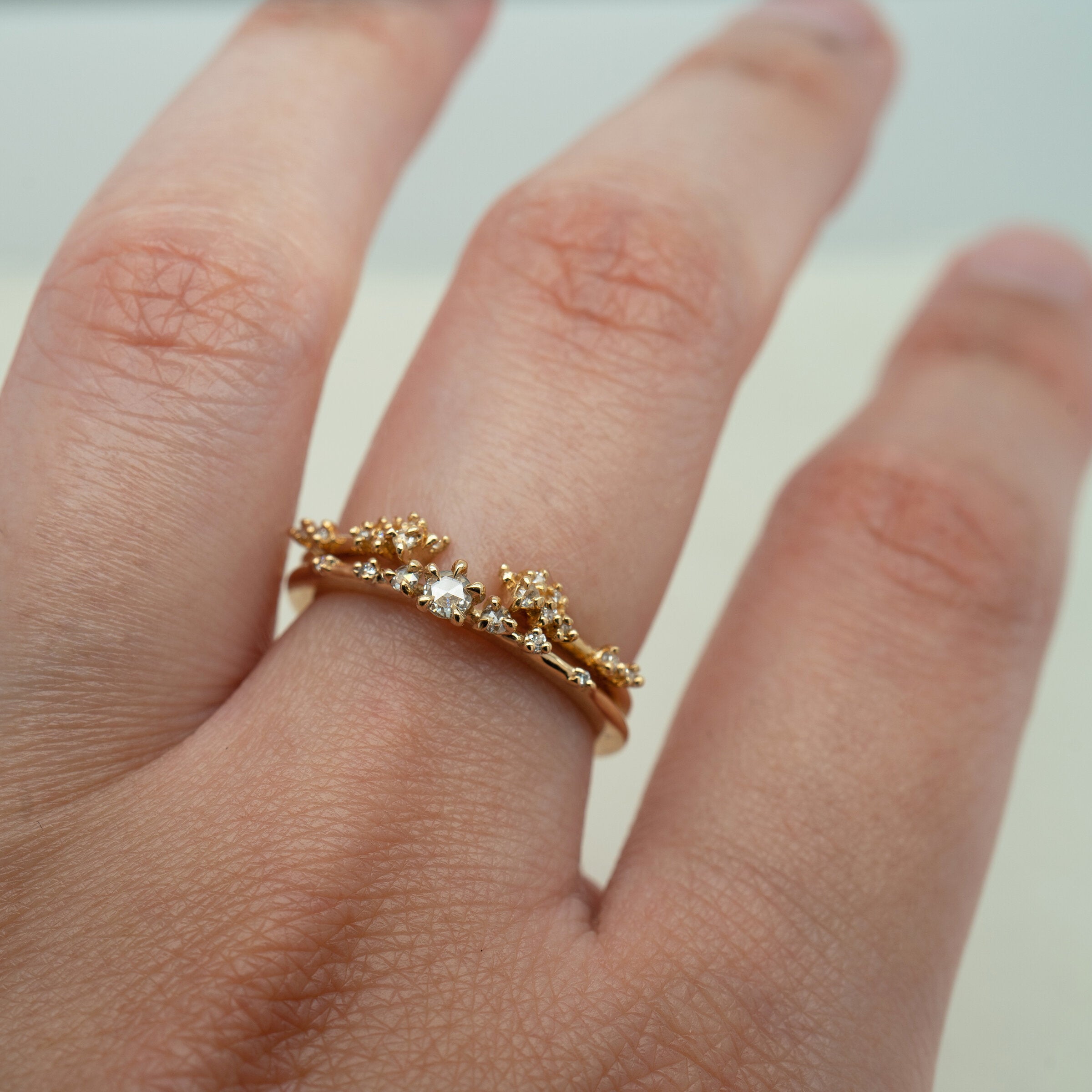 A stack of Laurie Fleming Jewellery rings worn on a hand. At the base of the finger is a "Water Lily Ring," a delicate solid gold stacking ring with round brilliant and rose cut diamonds scattered along the band. On top of that is a "Daphne Band", an open band that features clusters of both round brilliant and rose cut diamonds in varying sizes. The hand is in front of a pale grey background.
