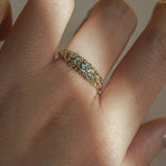 Laurie Fleming Jewellery's "Moss Signet Ring," a bold solid gold signet-style ring encrusted with varying shades and sizes of blue-green teal sapphires on the band. The ring is worn on a hand in front of a light grey background; most of the photo is in the shade, but a beam of light is shining perfectly across the ring.