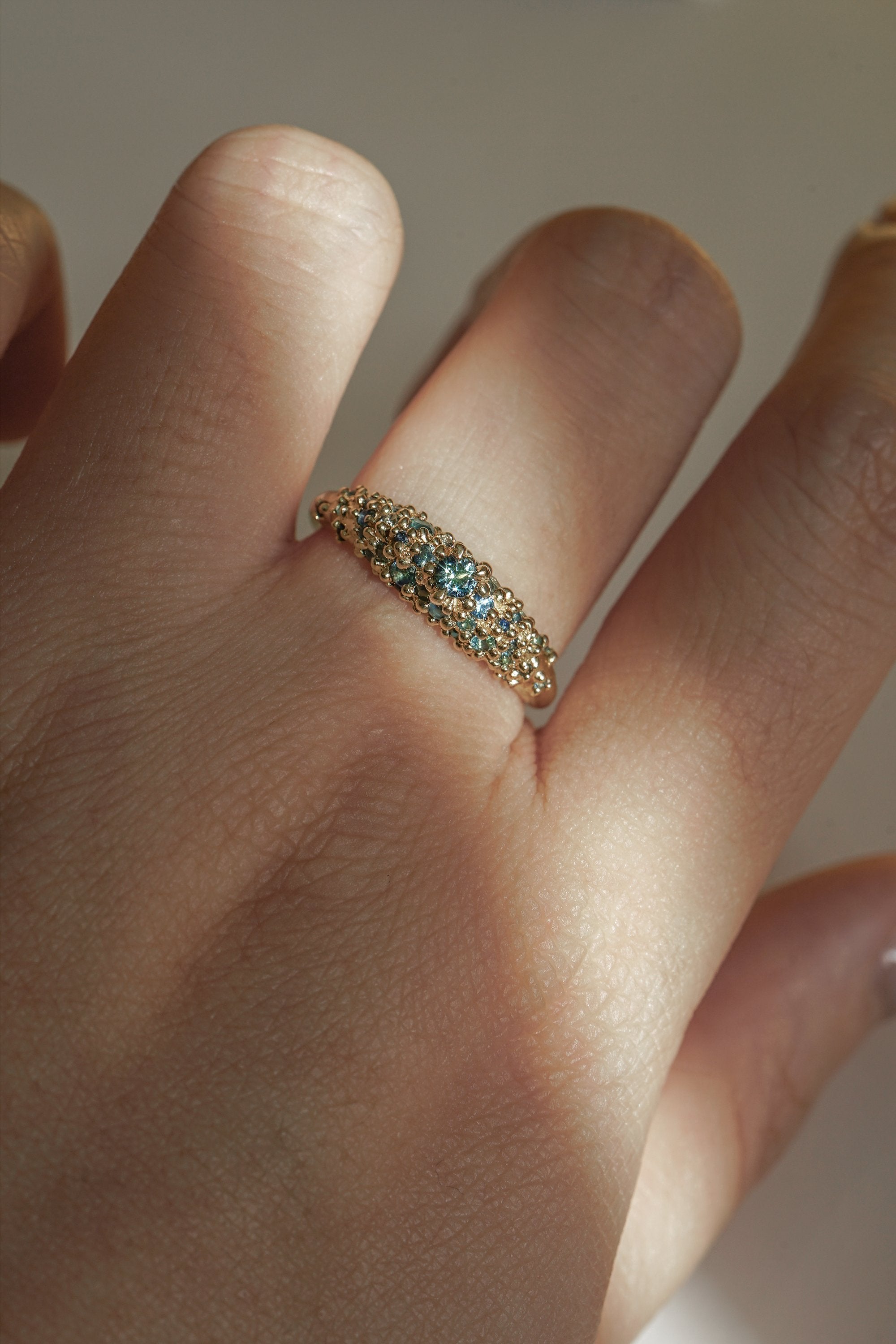 Laurie Fleming Jewellery's "Moss Signet Ring," a bold solid gold signet-style ring encrusted with varying shades and sizes of blue-green teal sapphires on the band. The ring is worn on a hand in front of a light grey background; most of the photo is in the shade, but a beam of light is shining perfectly across the ring.