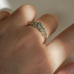 Laurie Fleming Jewellery's "Moss Signet Ring," a bold solid gold signet-style ring encrusted with varying shades and sizes of blue-green teal sapphires on the band. The ring is worn on a hand in front of a light grey background; most of the photo is in the shade, but a beam of light is shining perfectly across the ring.