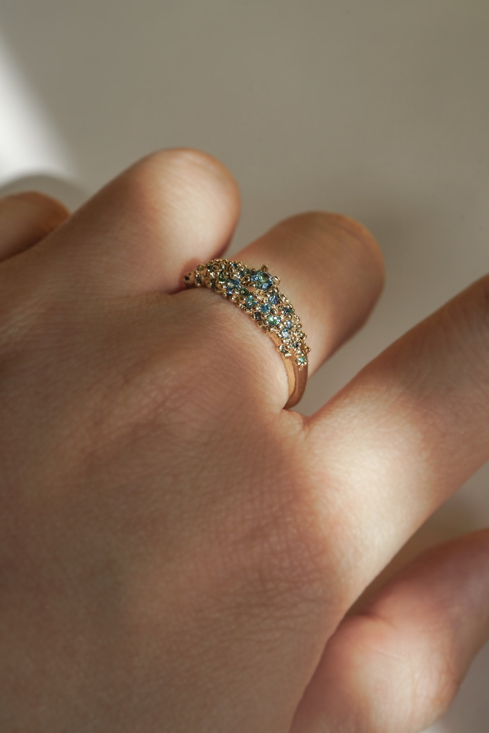 Laurie Fleming Jewellery's "Moss Signet Ring," a bold solid gold signet-style ring encrusted with varying shades and sizes of blue-green teal sapphires on the band. The ring is worn on a hand in front of a light grey background; most of the photo is in the shade, but a beam of light is shining perfectly across the ring.