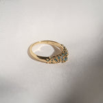 Laurie Fleming Jewellery's "Moss Signet Ring," a bold solid gold signet-style ring encrusted with varying shades and sizes of blue-green teal sapphires on the band. The ring is resting on smooth white fabric. There are some shadows cast over the frame, but the ring is in sunlight.
