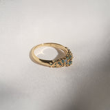Laurie Fleming Jewellery's "Moss Signet Ring," a bold solid gold signet-style ring encrusted with varying shades and sizes of blue-green teal sapphires on the band. The ring is resting on smooth white fabric. There are some shadows cast over the frame, but the ring is in sunlight.