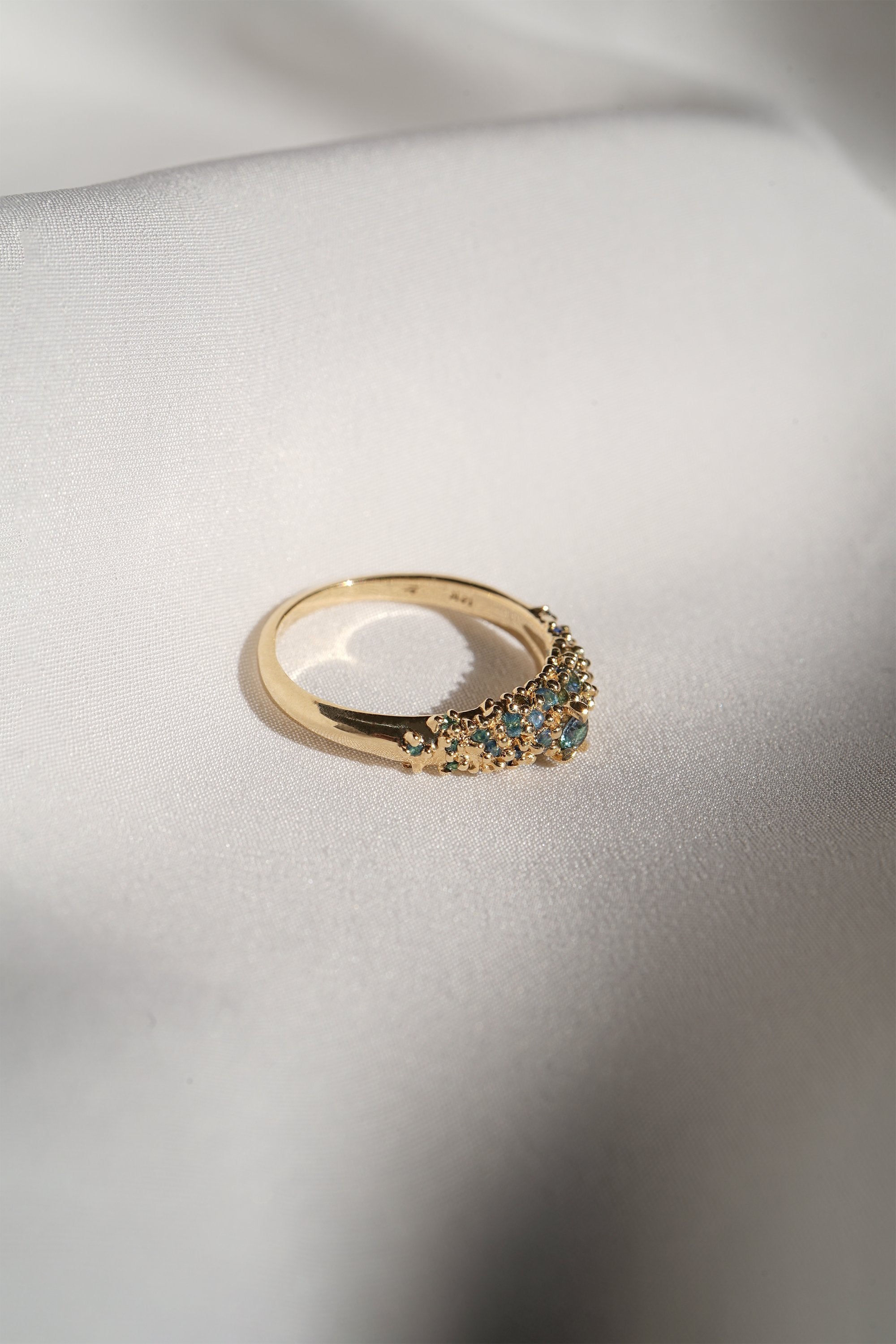 Laurie Fleming Jewellery's "Moss Signet Ring," a bold solid gold signet-style ring encrusted with varying shades and sizes of blue-green teal sapphires on the band. The ring is resting on smooth white fabric. There are some shadows cast over the frame, but the ring is in sunlight.