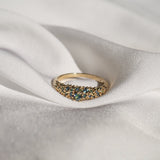 Laurie Fleming Jewellery's "Moss Signet Ring," a bold solid gold signet-style ring encrusted with varying shades and sizes of blue-green teal sapphires on the band. The ring is nestled in folds of smooth white fabric.
