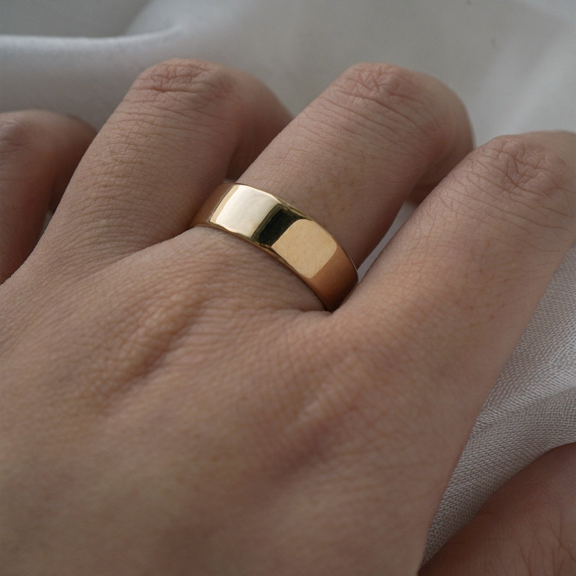 A stock photo of Laurie Fleming Jewellery's plain solid gold "Rowan" band, a 6mm wide flat comfort fit stacking ring/wedding band, handmade in Toronto. The ring is worn on a hand with a light grey background.