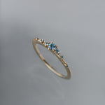 Laurie Fleming Jewellery's "Sapphire Water Lily Ring," a delicate solid gold stacking ring with blue-green teal sapphires scattered along the band. The ring is placed in front of a medium grey background.