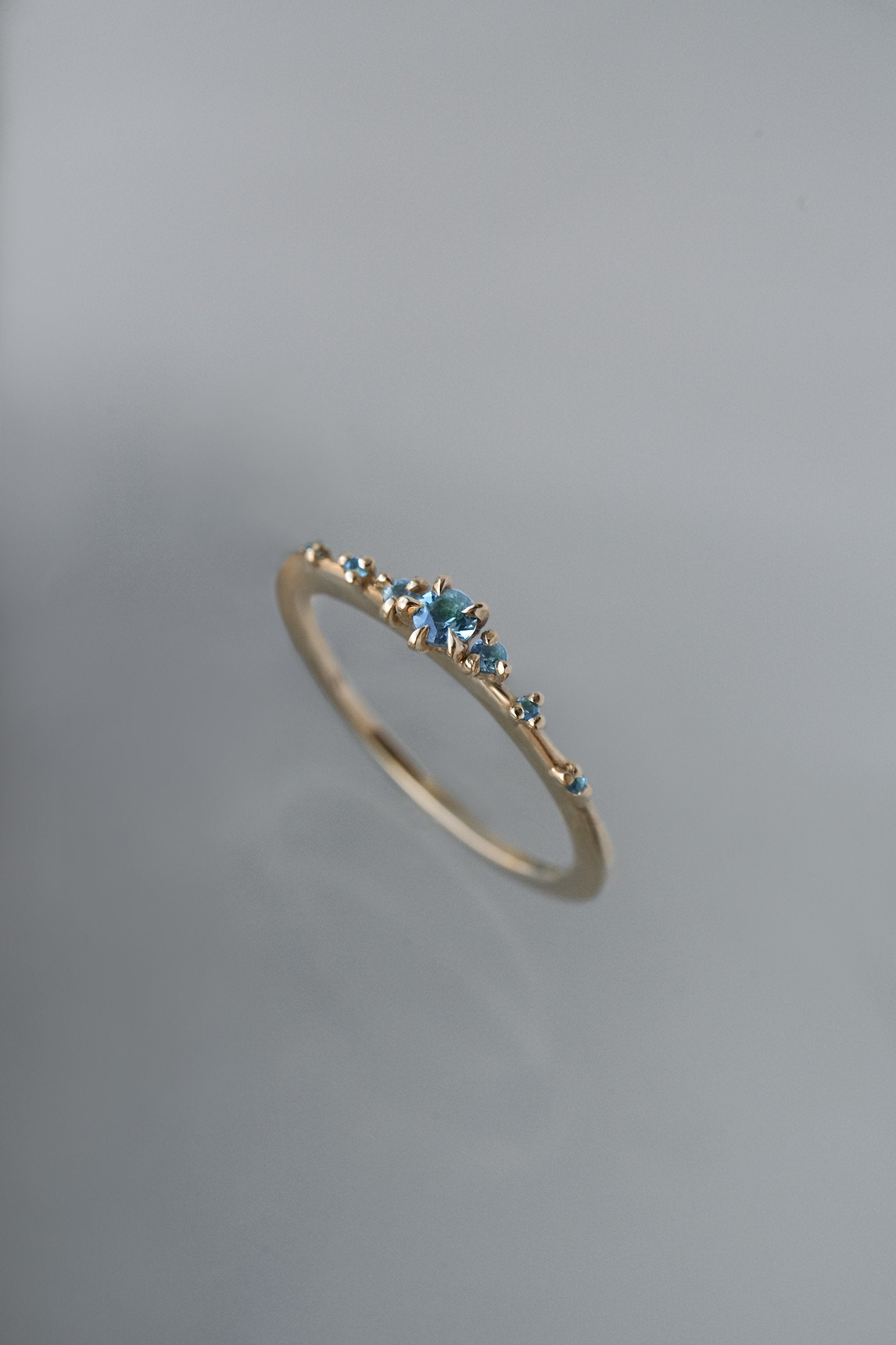 Laurie Fleming Jewellery's "Sapphire Water Lily Ring," a delicate solid gold stacking ring with blue-green teal sapphires scattered along the band. The ring is placed in front of a medium grey background.