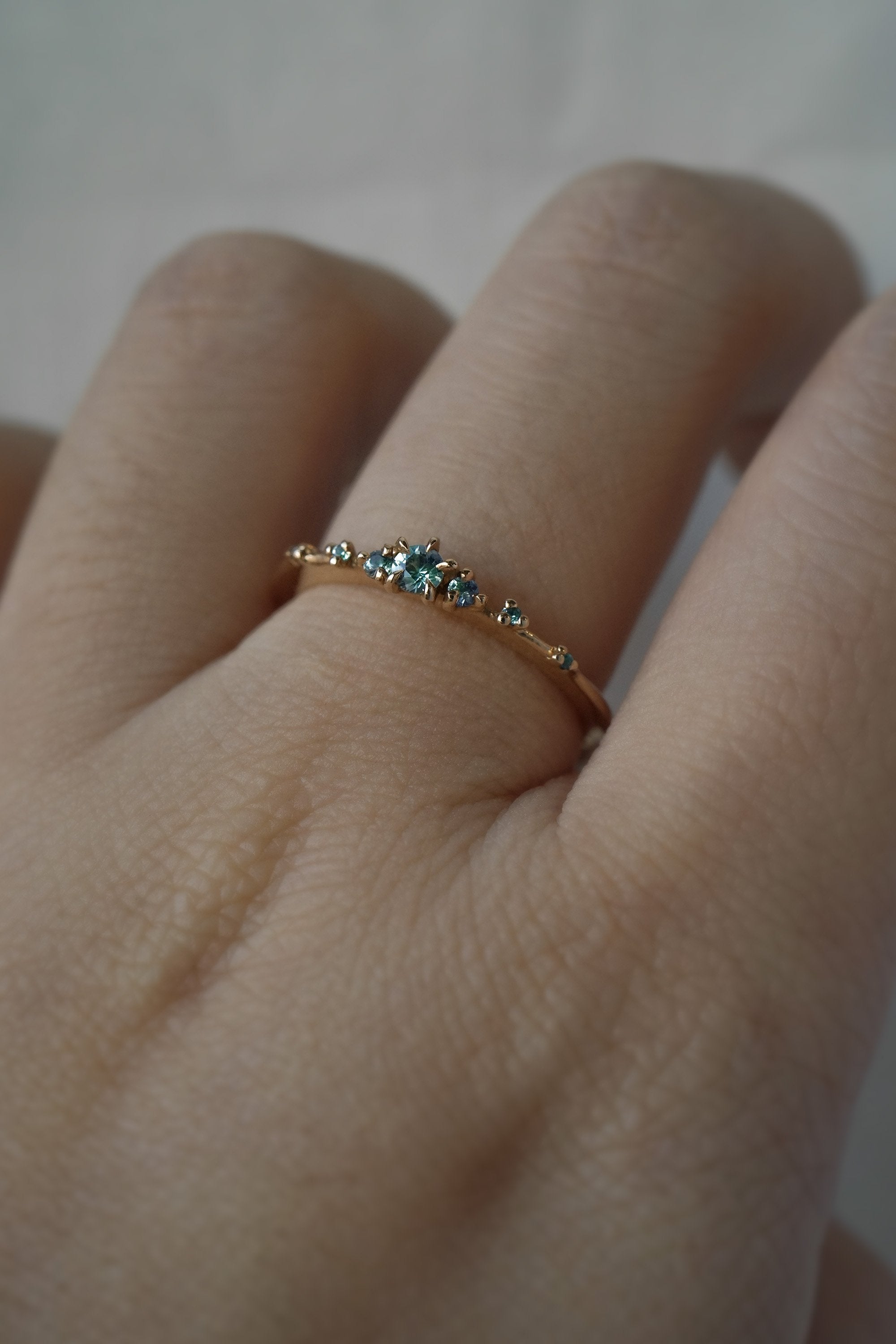 Laurie Fleming Jewellery's "Sapphire Water Lily Ring," a delicate solid gold stacking ring with blue-green teal sapphires scattered along the band. The ring is worn on a hand.